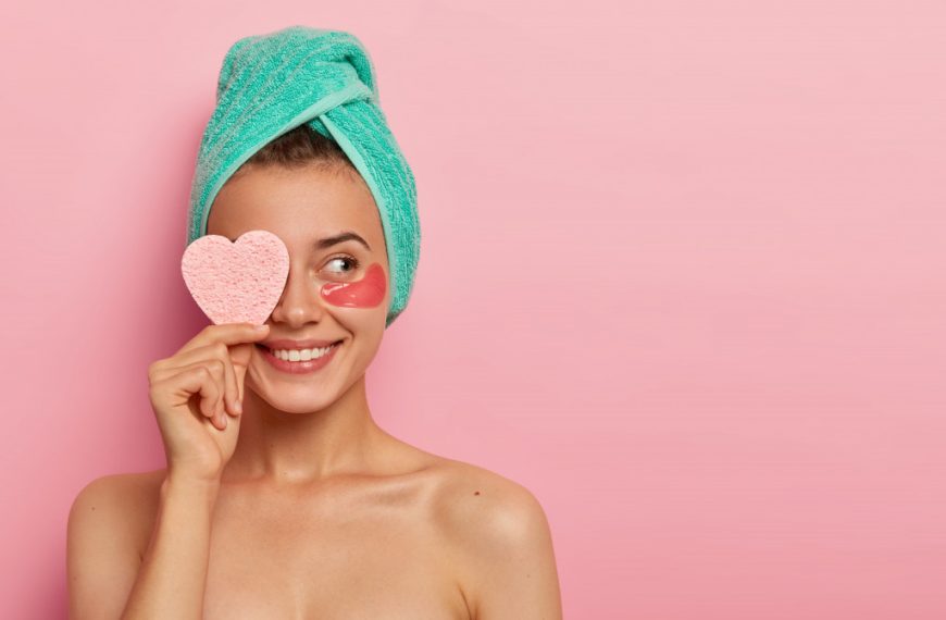 smiling woman holding heart-shaped pink sponge in face with towel in hair